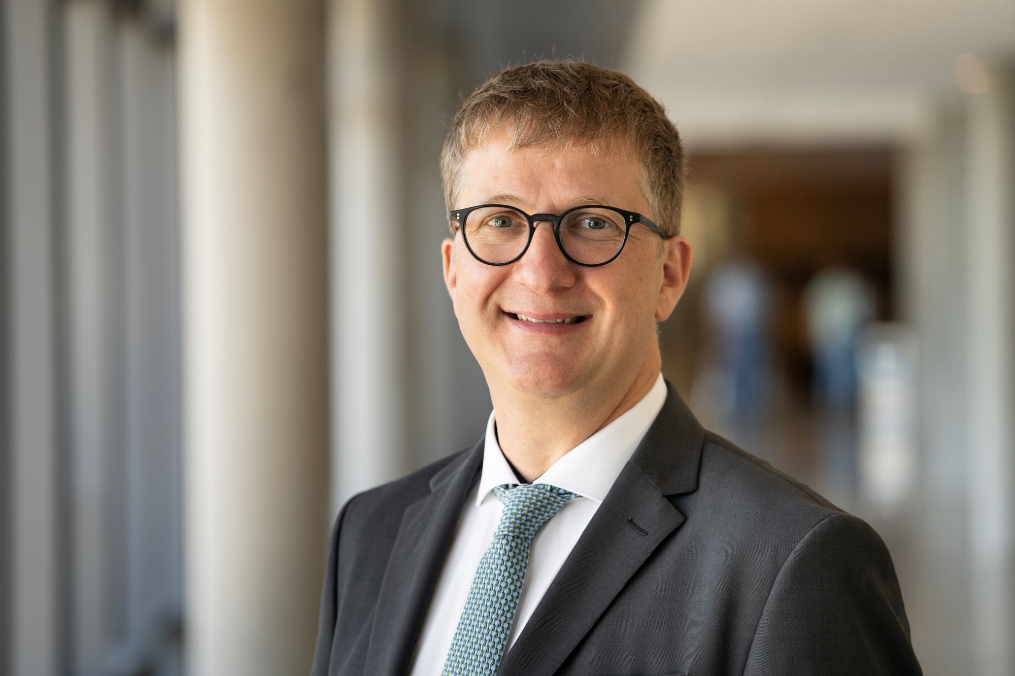 Sebastian Walther Porträtbild von Sebastian Walther im grauen Anzug und hellblauer Krawatte, dunkle Brille - im Hintergrund verschwommen ein Flur mit Säulen zu sehen.