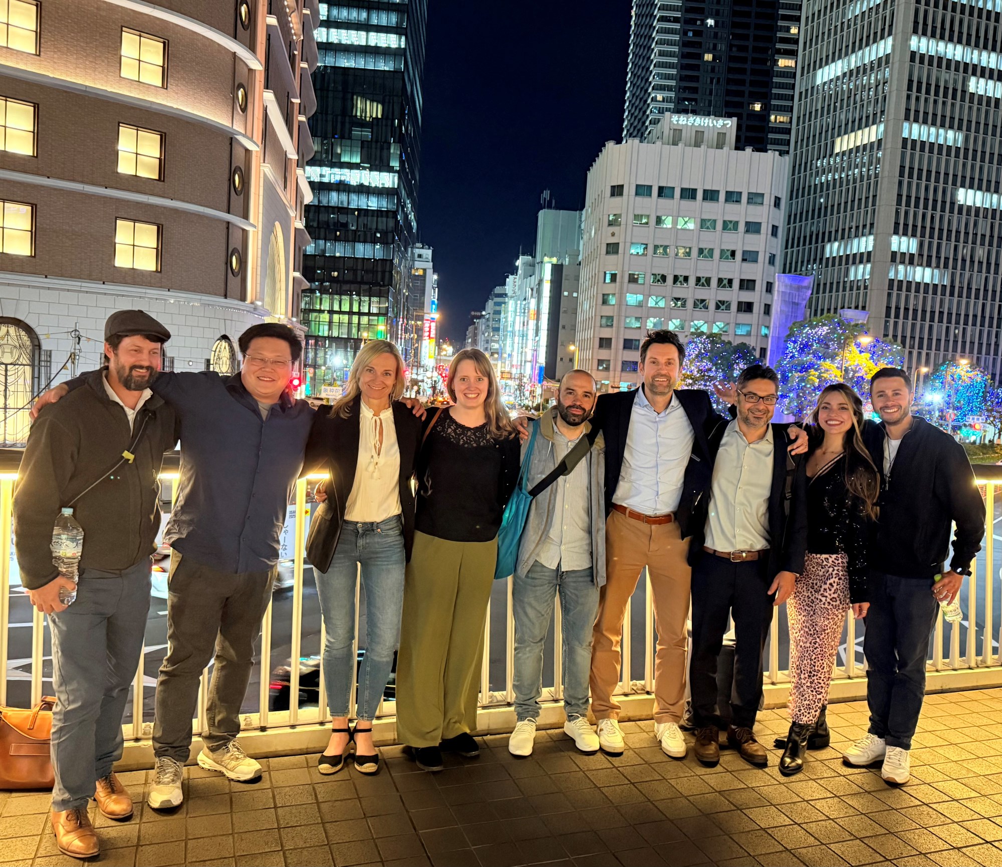 DZHI-Team in Osaka 9 Forschende stehen in Freizeitkleidung vor einem Geländer, im Hintergrund ist das Stadtbild von Osaka bei Nacht, Hochhäuser und bunt erleuchtete Straßen