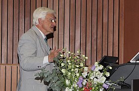 Klaus Viktor Toyka steht am Rednerpult im Hörsaal, vor ihm ein großer bunter Blumenstrauße in einer Bodenvase. 