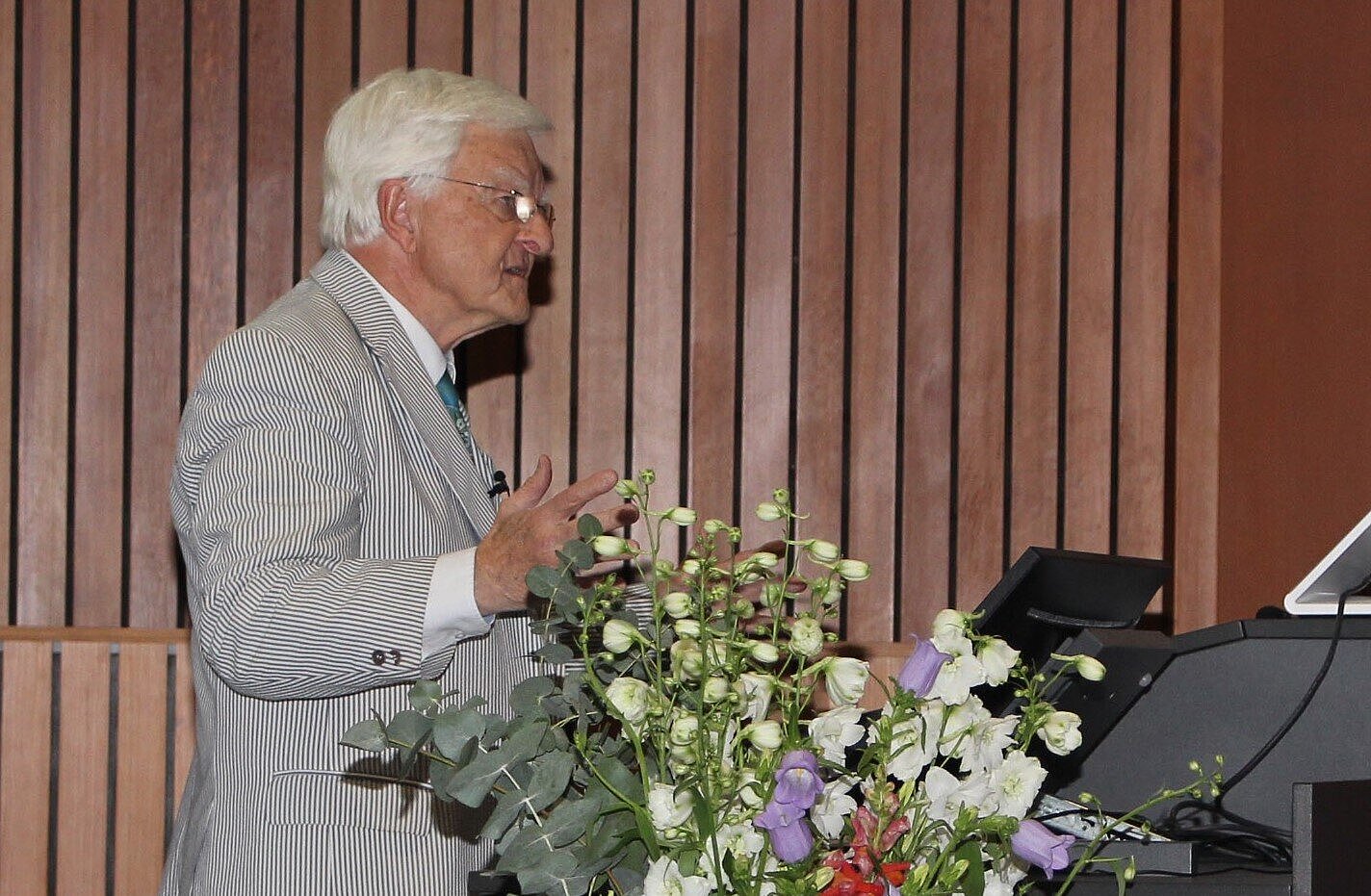 Klaus Viktor Toyka steht am Rednerpult im Hörsaal, vor ihm ein großer bunter Blumenstrauße in einer Bodenvase. 