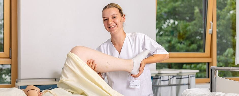 zu den Ausbildungen Physiotherapie, Massage Symbolbild: Azubine aus dem Bereich Physiotherapie am Bett einer Patientin. Die Azubine hält das Bein der Patientin und führt mit ihr Übungen aus.