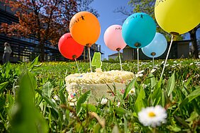 Torte mit bunten Smarties und mit einer grünen Kerze in 4-Form auf einer Wiese, drumherum bunte MIAI-Luftballons 