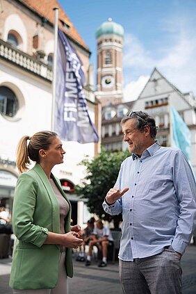 Bayern Gesundheitsministerin Judith Gerlach bei einem Gespräch mit einem Mann. Im Hintergrund sieht man eine Fahne und Münchens Kirche.