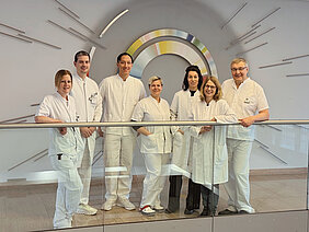 Das Team aus Endokrinologie und Nuklearmedizin steht in weißer Arbeitskluft nebeneinander im Flur des Zentrums für Innere Medizin hinter einem Treppengeländer, vor einer Wanddekoration der NUK