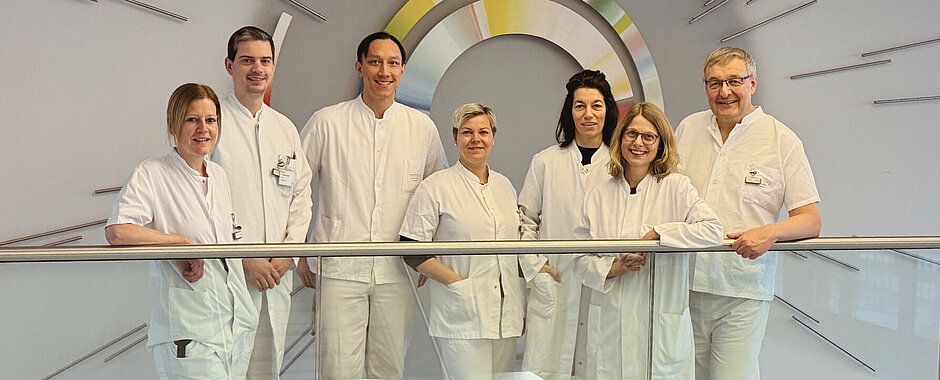 Das Team aus Endokrinologie und Nuklearmedizin steht in weißer Arbeitskluft nebeneinander im Flur des Zentrums für Innere Medizin hinter einem Treppengeländer, vor einer Wanddekoration der NUK
