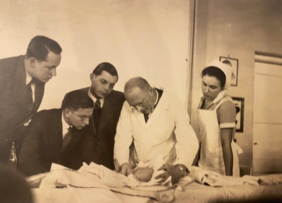 Das Bild zeigt Hans Rietschel, der im weißen Kittel einen Säugling untersucht, daneben eine SChwester in Schwestertracht, links schauen drei Studierende in dunklen Anzügen zu. 