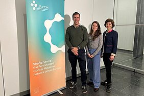 Die drei Forschenden stehen neben einem Roll-up der Joint Action, auf dem der Slogan steht "Strengthening the Personalised Cancer Mecicine Network across Europe 