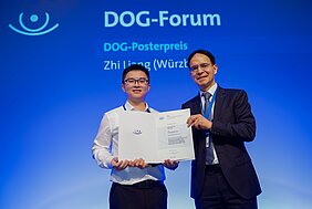 Professor Hattenbach und Zhi Liang stehen auf der Bühne, DOG-Forum steht auf blauem Hintergrund, beide halten Urkunde in den Händen 