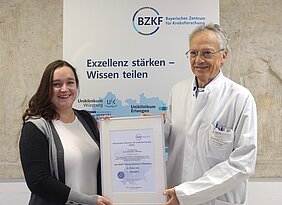Die Preisträgerin Julia Krug und Ralf Bargou halten die eingerahmte Urkunde in den Händen, sie stehen vor einem Roll-up des BZKF mit den Worten Exzellenz stärken - Wissen teilen. 