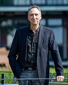 Prof. Dr. Meining steht schmunzelnd hinter einem Geländer an welchem er sich mit seiner linken Hand festhält. Im Hintergrund kann man eine Wiese und ein verglastes Gebäude, welches aber verschwommen dargestellt wird, erkennen.