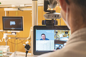 Hintere Kopfansicht einer Person, die auf einen Monitor schaut ,auf dem ein Arzt vom UKW zu ihr spricht. In weiteren Feldern auf dem Monitor sind das Patientenbett und Parameter zu sehen.   