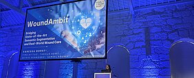 App Wunderkint - Präsentation Die Informatikerin Vanessa Borst auf der Bühne am Rednerpult, im Hintergrund das Startbild ihrer Präsentation auf einem riesigen Monitor. Der Konferenzsaal ist in blauem Licht.