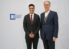 Ugur Uslu und Paul Pauli posieren in Anzügen nebeneinander vor einer Wand mit Logo der Universität Würzburg 