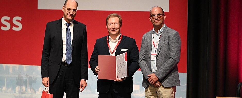 Die drei Herren stehen in Anzügen auf der Kongressbühne, Preisträger Patrick-Pascal Strunz hält eine Urkunde in den Händen.