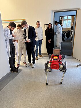 Roboterhund HELIA steht vor dem Eingang zur Bibliothek in der Hautklinik. Außenherum stehen einige Zuschauende und einige Personen, die einen Fragebogen ausfüllen, darunter Mitarbeitende der Hautklinik und Forschende.