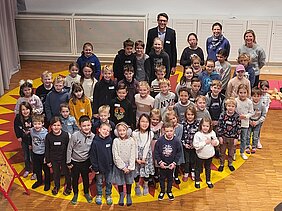 Das Bild wurde von oben runter auf eine Schar von Kindern fotografiert, die auf einem runden gelben Teppich mit roten Zacken stehen. Im Hintergrund der Kinder stehen der Ärztliche Direktor und zwei Resilienztrainerinnen. 