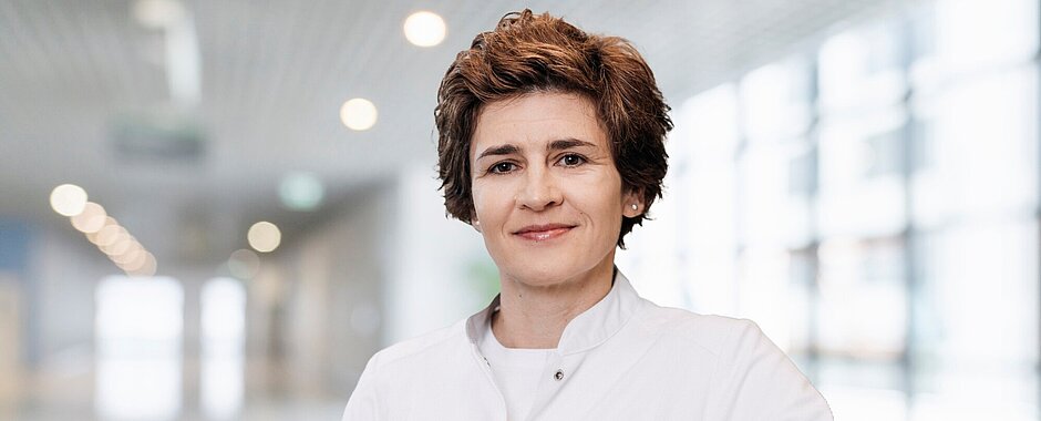 Anke Bergmann steht in weißem Kittel in einem Flur, der Hintergrund ist sehr unscharf. Das Bild ist im Querformat, Frau Bergmann ist bis zum Bauchnabel zu sehen. 