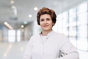 Anke Bergmann steht in weißem Kittel in einem Flur, der Hintergrund ist sehr unscharf. Das Bild ist im Querformat, Frau Bergmann ist bis zum Bauchnabel zu sehen. 