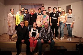 Auf dem Foto sieht man mehrere Personen, die teilweise verkleidet sind und auf einer Bühne stehen bzw. sitzen. 