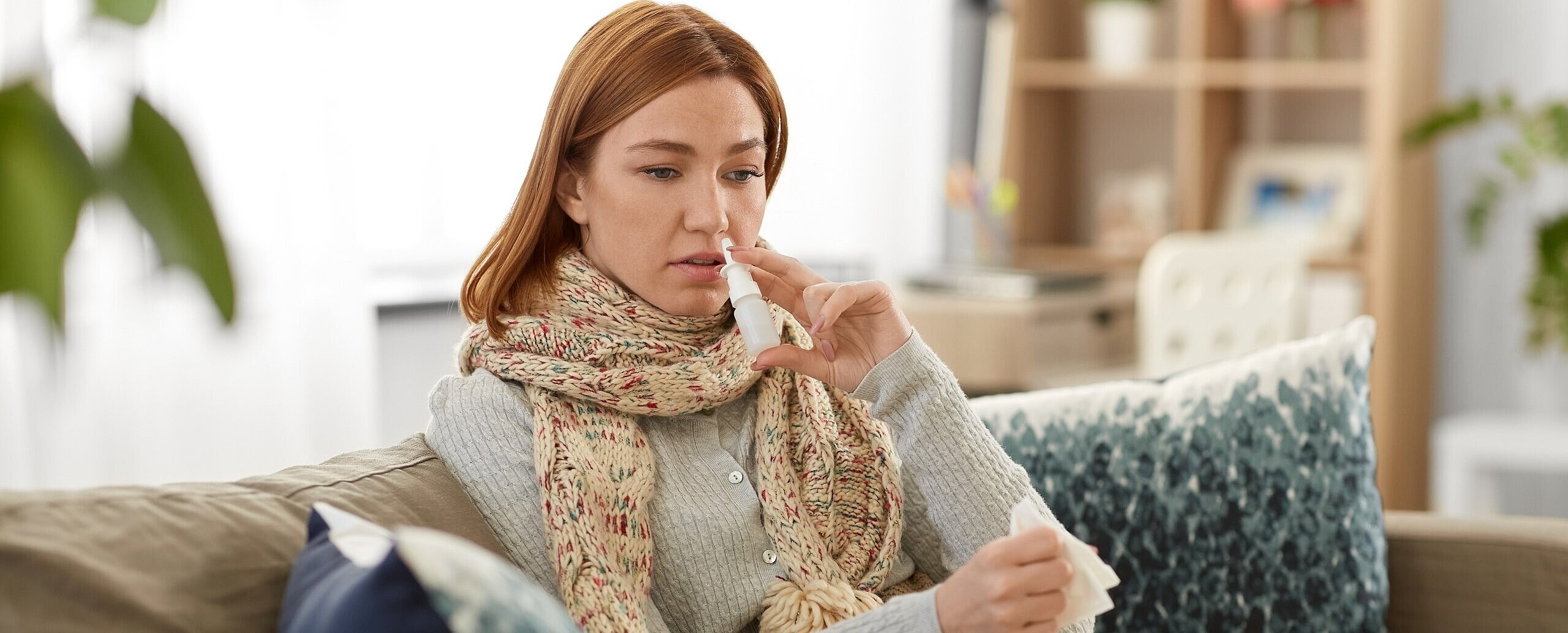 Frau mit Schal und Decke sitzt auf einem Sofa - mit einem Taschentuch in der einen Hand und einem Nasenspray in der anderen Hand. Das Nasenspray führt sie gerade zur Nase. 