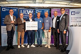 Sieben Männer stehen auf der Bühne der MS Wissenschaft, in der Mitte bzw. dritter von links Michael Hudecek mit Scheck in der Hand, im Hintergrund Roll-ups. 
