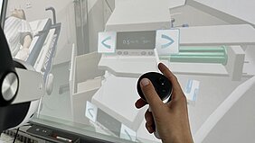 VR-Situation aus Sicht des Anwenders: links im Bildrand die VR-Brille und in der Mitte des Bildes die Hand des Anwenders mit VR-Gerät - im Hintergrund die virtuelle Welt mit Patient und Geräten