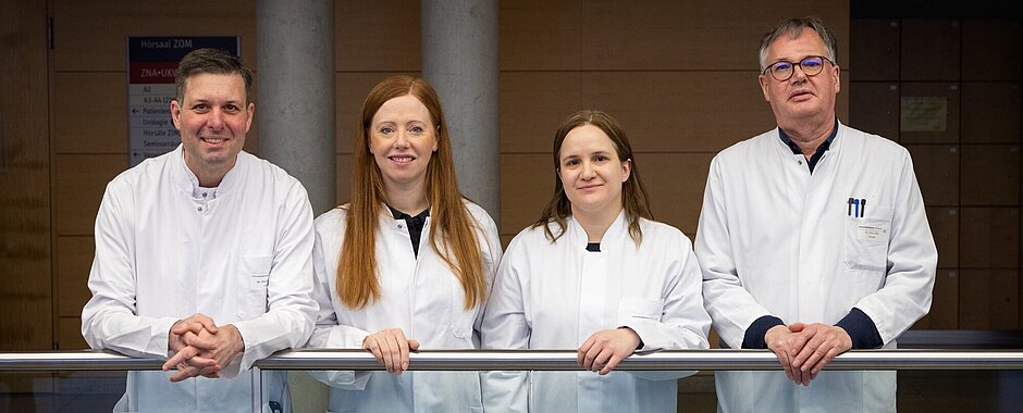 Die vier Forschenden stehen in weißen Laborkitteln an einem gläsernen Geländer und stützen sich ab. 