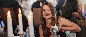 Foto zeigt Vanessa Borst, die einen Pokal in der Hand hält. Im Hintergrund sitz verschwommen verschiedene Leute an Tischen. Neben der Person sieht man auch verschwommen fünf Kerzen brennen.