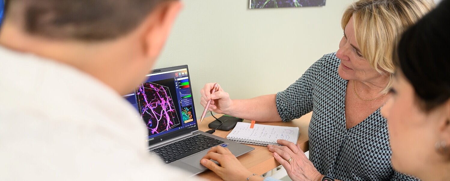 Mateo Andrade Mier, Carmen Villmann und Esra Türker sitzen vor einem Laptop und betrachten ein mikroskopisches Bild, Mateo ist nur von hinten zu sehen. 