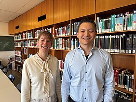 Die Wissenschaftlerin Lisa Harder-Rauschenberger steht mit hochgesteckten Haaren, in heller Schleifenbluse und heller Strickjacke neben Chi Wang Ip im hellblauen Hemd vor dem Bücherregal in der Bibliothek der Neurologie. 