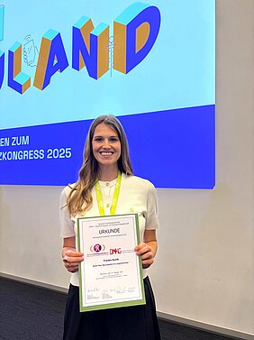 Franka Kunik  Fanka Kunik steht vor der Bühne des Deutschen Schmerzkongresses und hält die Urkunde in den Händen, im Hintergrund die Leinwand mit einer Folie, auf der Neuland steht.