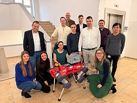 Die Forschenden aus den verschiedenen Zentren stehen im Treppenhaus der Dermatologie, vor ihnen ist der rote Roboter-Hund Helia. Einige Personen knien neben HELIA.