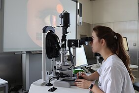 Studierende sitzt am Simulator für die Spaltlampe - der Patient ist ein schwarzes Kopfmodel. Im Hintergrund ist das Auge in groß auf einem Monitor zu sehen. 
