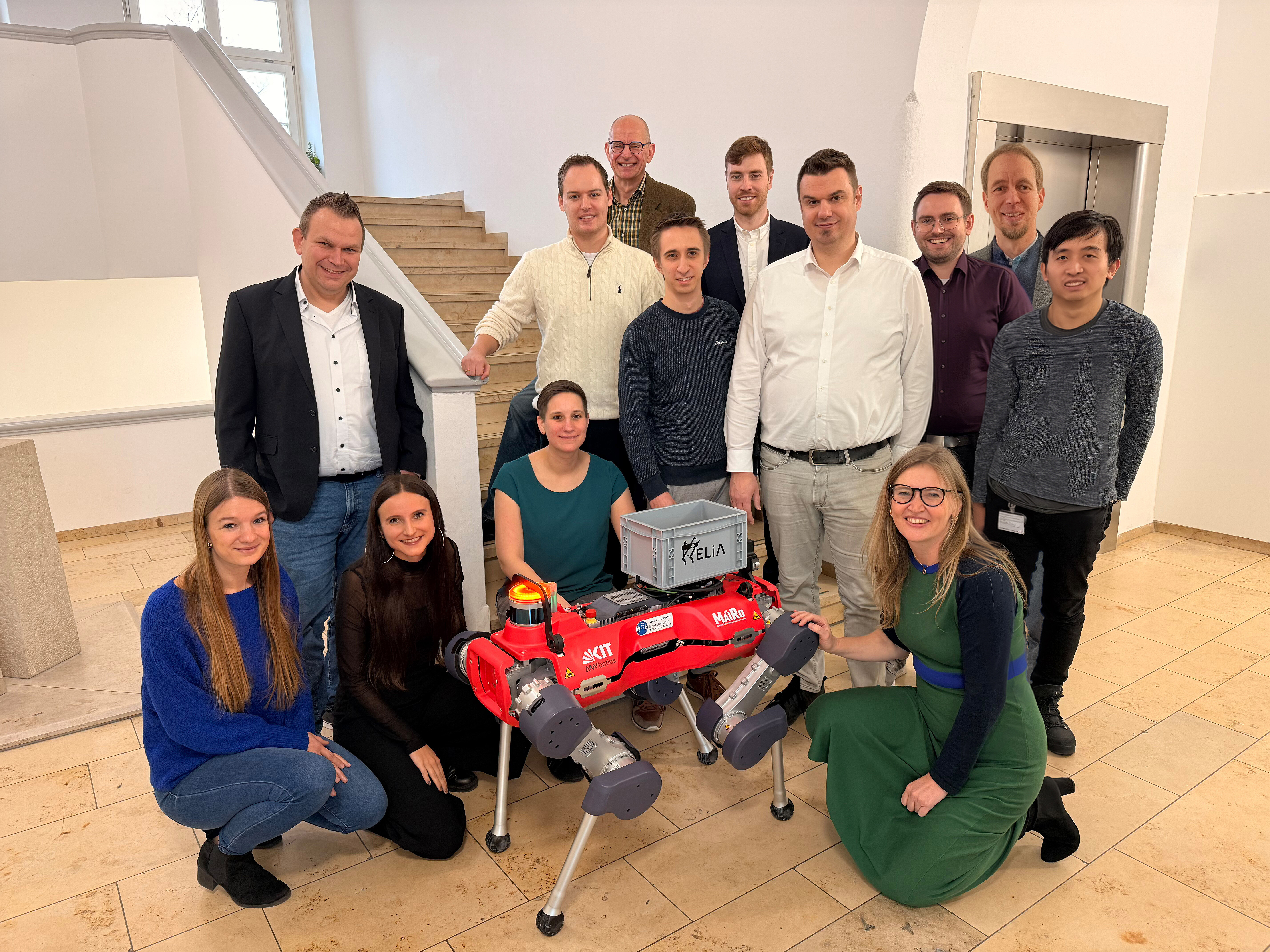 Gruppenfoto des HELIA-Teams mit Roboterhund. Fotografin: Kim Sammet Gruppenfoto des HELIA-Teams im Treppenhaus der Dermatologie mit dem roten Roboterhund HELIA in der Mitte. Einige der Personen knien neben dem Roboter.