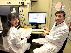 Die beiden Forschenden sitzen in weißen Kitteln im Labor vor einem Rechner und schauen in die Kamera. 