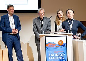 Bild zeigt Herrn Prof. Lock, Herrn Prof. Lopau, Frau Dr. Wagner und Frau Dr. Herzog. Alle vier stehen an einem Pult und zeigen eine Präsentation. 