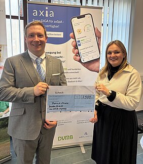 Der Gewinner und die Spenderin halten einen auf DinA2 ausgedruckten Scheck hoch, dahinter ein Rollup von der App Axia.  