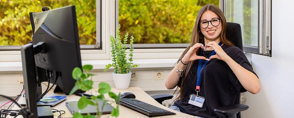 Zu den Ausbildungen in Bereichen Bürokommunikation und Gesundheitswesen Symbolbild: Azubine am Schreibtisch. Mit den Händen formt sie ein Herz.