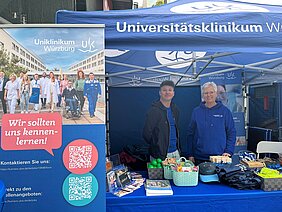Unter einem UKW-Pavillon stehen vor einem Give-Away Tisch zwei Mitarbeiter. Links steht ein Roll-Up "Wir sollten uns kennen lernen".