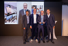 Hermann Einsele steht mit Preis und Kollegen vor einer Leinwand mit Bild, das ihn würdigt. 