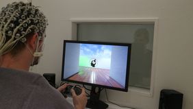 Ein Proband sitzt vor einem Monitor und hat eine Haube für das Elektroenzephalogramm auf dem Kopf