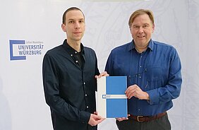 Die beiden Herren stehen vor einer Wand mit Logo der Uni Würzburg und halten die Ernennungsurkunde in den Händen 