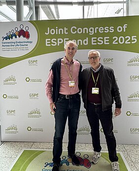 Die beiden Wissenschaftler posieren vor einer Stellwand beim Kongress der ESPE und ESE mit Logos 