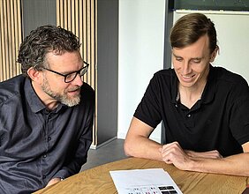 Die beiden Wissenschaftler sitzen am Tisch - vor sich der Ausdruck einer Publikation 
