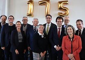 Auf dem Personen die sich zu einem Gruppenbild zusammengestellt haben. Im Hintergrund sind drei goldene Luftballons als Zahl 175 zu erkennen.