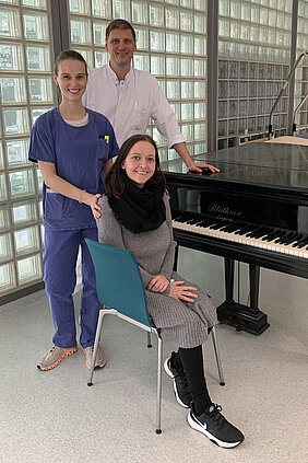 Auf dem Bild sind drei Personen zu sehen. Im Hintergrund ist eine Glassteinwand zu sehen. Die Ärztin hat blaue OP-Kleidung an und hat ihre Hände auf den Schultern der Patientin. Der Prof. hat einen weißen Kittel an und stützt sich mit einer Hand auf einem Klavier ab. Die Patienten sitzt auf einem Stuhl vor dem Klavier.