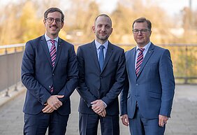 Charis Kalogirou, Marcel Schwinger und Christian Thomas stehen auf einer Terrasse nebeneinander und posieren in Anzug und Krawatte für den Fotografen.