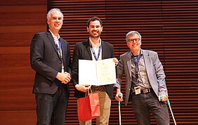 Preisträger Dr. Mario Detomas mit Urkunde auf der Bühne, rechts steht  Prof. Jan P. Tuckermann auf Krücken, links von ihm Prof. Martin Fassnacht.