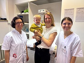 Die Mutter Hannah Hinz hält im Untersuchungsraum ihren Sohn auf dem Arm, daneben steht das Studienteam in weißer Kluft. 