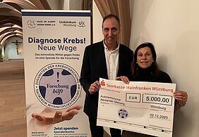Foto zeigt Spendenübergabe. Prof. Meining und Frau Nelkenstock halten einen Scheck über 5.000 Euro in der Hand. Links im Hintergrund steht ein Roll-up mit Diagnose Krebs - Neue Wege mit dem Logo Forschung hilft.